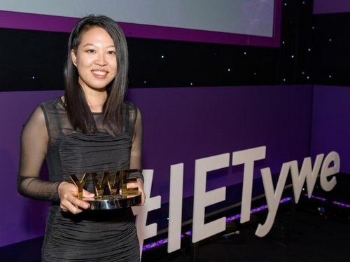 Ying Wan Loh wearing a black evening dress, holding her gold award. She is in front of the a display with the words #IETywe. Ying is a young Asian woman with shoulder length hair and an undercut. Her dress is black with see-through sleeves.