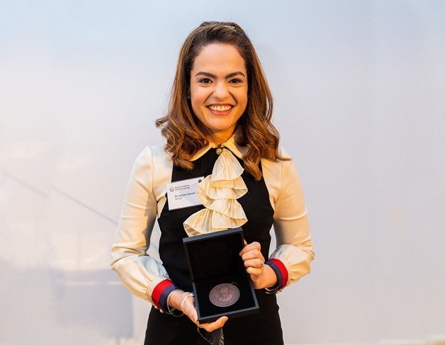 A smiling young Latina woman holding a bronze medal in a presentation box. She has curled shoulder-length brown hair, and is wearing a ruffled white shirt and dark pinafore dress.