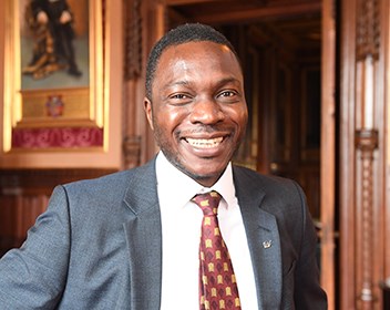 Photograph of Ugbana Oyet. A smilling black man with a short beard, photographed in a room with wood panelling, red velvet wallpaper and a gold-framed portrait in the background. He is wearing a grey suit, a tie with the House of Commons crest, and an IET lapel pin. Image: ©UK Parliament/Jessica Taylor https://creativecommons.org/licenses/by/3.0/