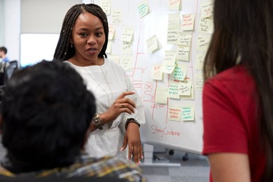 photo of three people in front of a whiteboard covered in post-it notes. We can see the backs of two figures, and frames is a black woman with braids, wearing a white gathered shirt