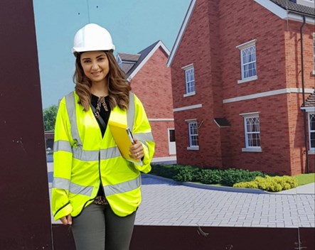Tia wearing a hard hat and hi vis jacket. She has long curled hair and is holding a clipboard