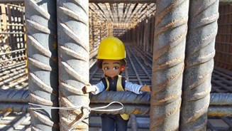 Lottie doll in hard hat on building site around huge steel beams