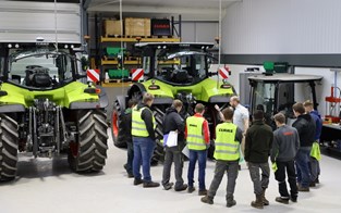 nine students around a speaker. They are in front of two huge mechanical vehicles. Some people are wearing hi-vis vests