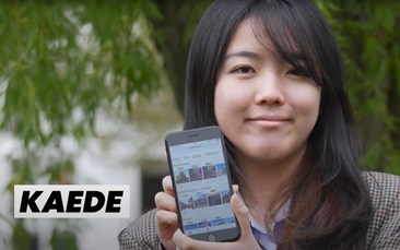 Smiling East Asian girl with long dark hair and a fringe. She is holding up a smartphone showing her new app. She is wearing a tweed jacket and there is a tree out of focus in the backgorund