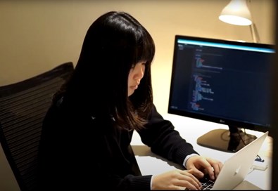 Kaede sat at a laptop. To one side is a computer monitor showing coding.