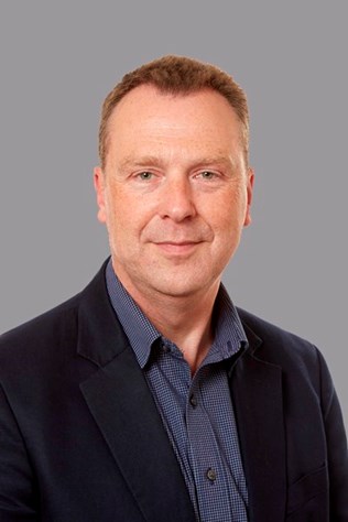 Portrait photo of John Chudley, a middle-aged caucasian man with short red hair. He is wearing a blue buttoned shirt and a dark suit jacket.