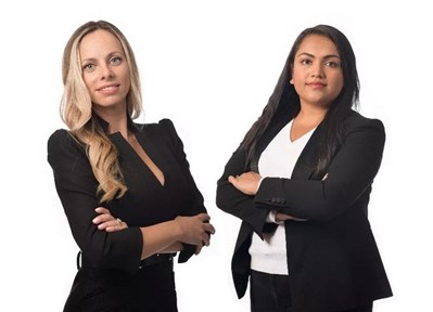 Photo of two young women wearing smart business clothes, with their arms folded, smiling at the camera. They are Eftychia Koursari (left) and Nipuni Karunaratne (right)