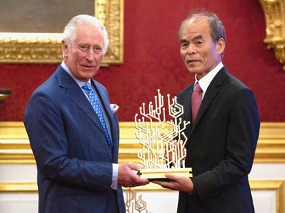 Prince Charles giving a gold trophy to Shuji Nakamara. Both men are wearing suits & ties. The trophy is an elaborate gold pattern, it looks like a computer motherboard.