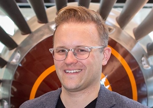 Headshot of Dr Mark McBride-Wright: a caucasian man with glasses & sandy hair in a short quiff. He is wearing a grey suit jacket and stood in front of a large, circular piece of engineering