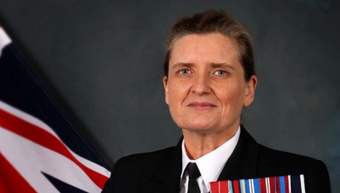 Captain Jo Deakin OBE CEng FIMechE RN, in uniform and wearing medals, next to a British flag