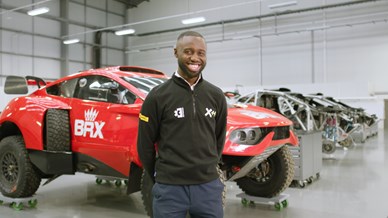 Engineer (black, male, short hair, black sweatshirt, smiling) standing in front of a red motorsport card